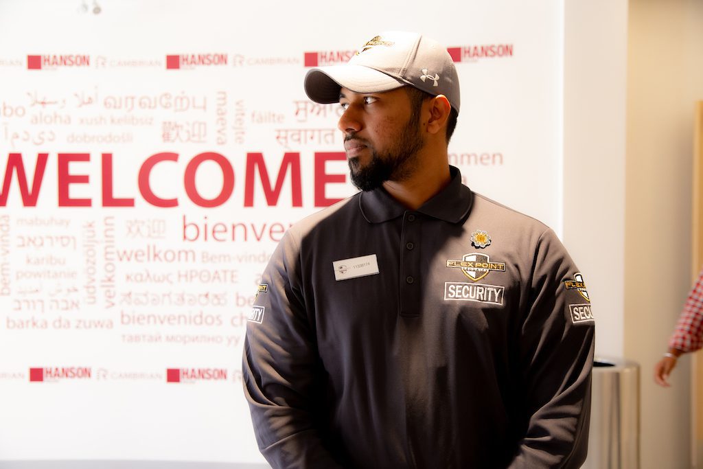 A Flex Point Security guard standing in front of a large sign that says the word "welcome" in many languages