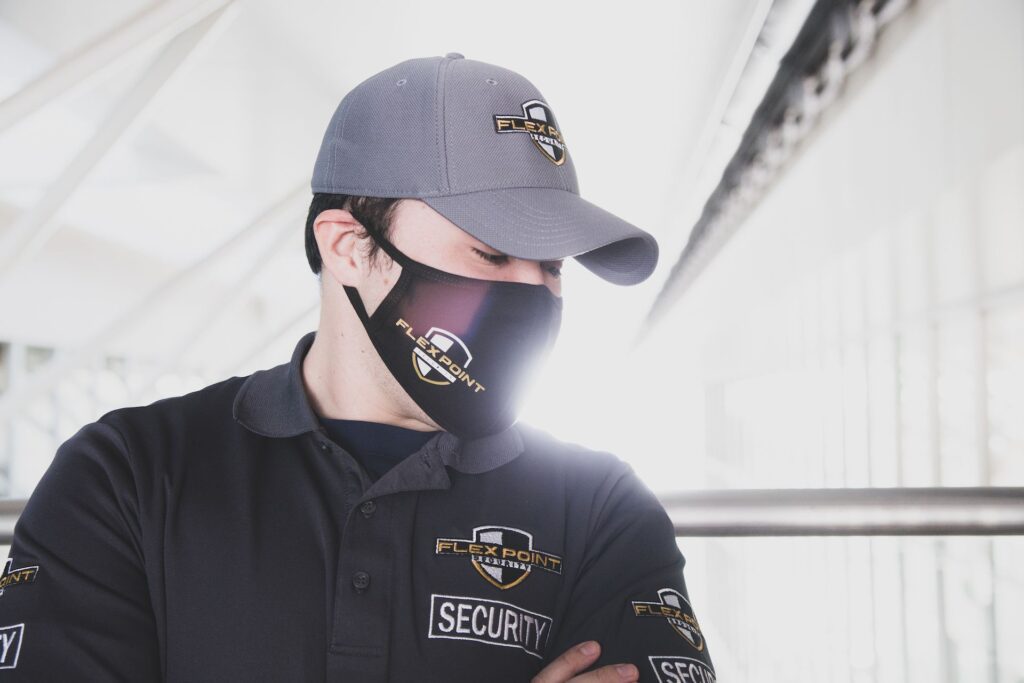 A Flex Point Security guard wearing a branded hat, shirt and mask with arms folded and looking down while on-site