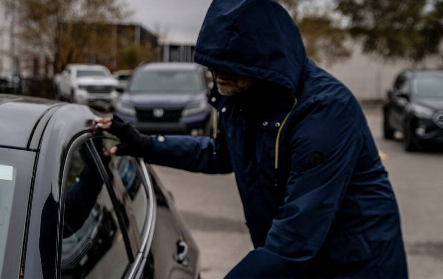 A criminal dressed in dark clothing with their face covered trying to break into a vehicle